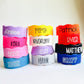 Colorful personalized silicone bottle bands stacked in three columns on a white background. The stretchy reusable labels come in bright colors including pink, purple, yellow, red, blue, white, and black, each customized with children’s names in various font styles and filled with permanent silicone ink. These custom silicone cup and bottle labels fit baby bottles, water bottles, sippy cups, and condiment bottles, making them ideal for daycare, school, and everyday organization.