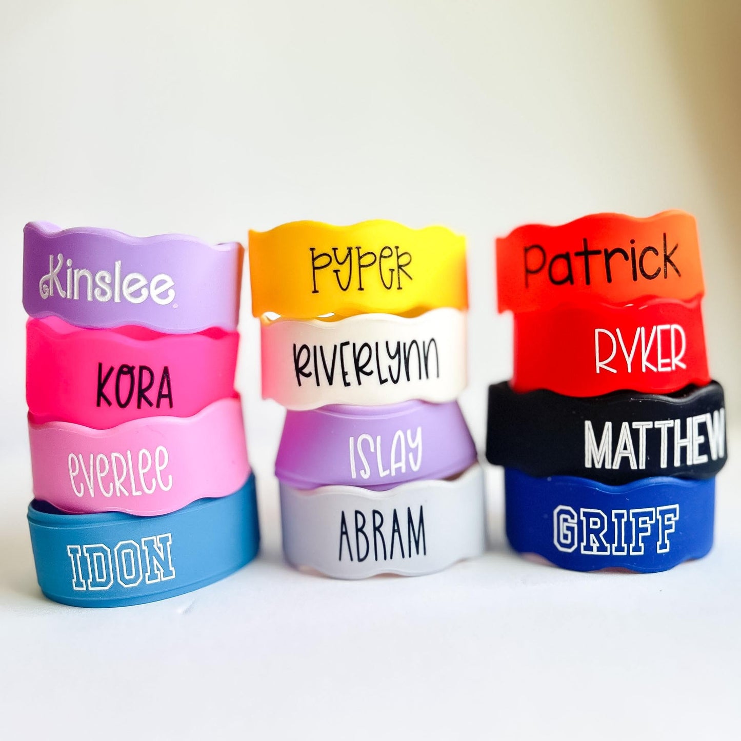 Colorful personalized silicone bottle bands stacked in three columns on a white background. The stretchy reusable labels come in bright colors including pink, purple, yellow, red, blue, white, and black, each customized with children’s names in various font styles and filled with permanent silicone ink. These custom silicone cup and bottle labels fit baby bottles, water bottles, sippy cups, and condiment bottles, making them ideal for daycare, school, and everyday organization.