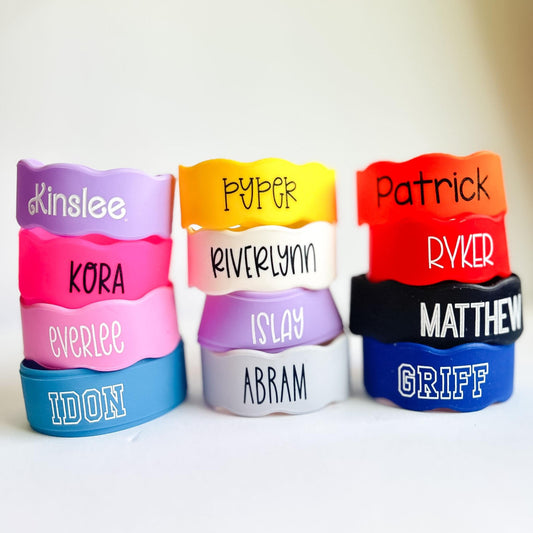 Colorful personalized silicone bottle bands stacked in three columns on a white background. The stretchy reusable labels come in bright colors including pink, purple, yellow, red, blue, white, and black, each customized with children’s names in various font styles and filled with permanent silicone ink. These custom silicone cup and bottle labels fit baby bottles, water bottles, sippy cups, and condiment bottles, making them ideal for daycare, school, and everyday organization.
